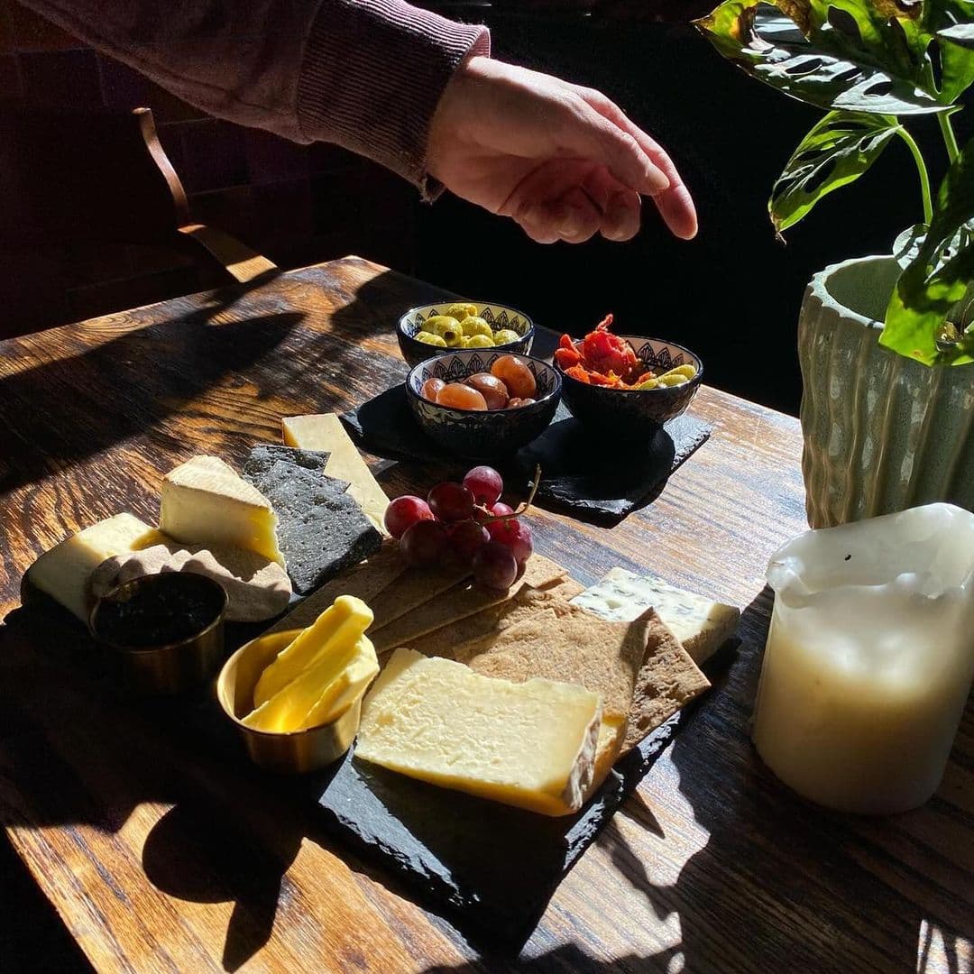 Holding Patterns cheese platter gourmet snacks dining experience A beautifully arranged cheese platter featuring various types of cheese, crackers, and fresh grapes, set on a wooden table. Small bowls of olives and dried fruits are also present, along with a lit candle and a green plant in the background. The scene is illuminated by natural sunlight, creating a warm and inviting atmosphere. **Location: Holding Patterns**