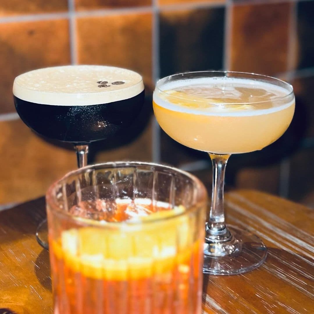 Holding Patterns cocktails experience nightlife tourism Two elegant cocktails sit on a wooden table at Holding Patterns. One is a dark drink topped with foam and coffee beans, while the other is a light, citrusy cocktail in a coupe glass. A third drink in a textured glass, featuring a vibrant orange hue and a slice of orange, is partially visible in the foreground. The background has warm-toned tiles, creating a cozy atmosphere.