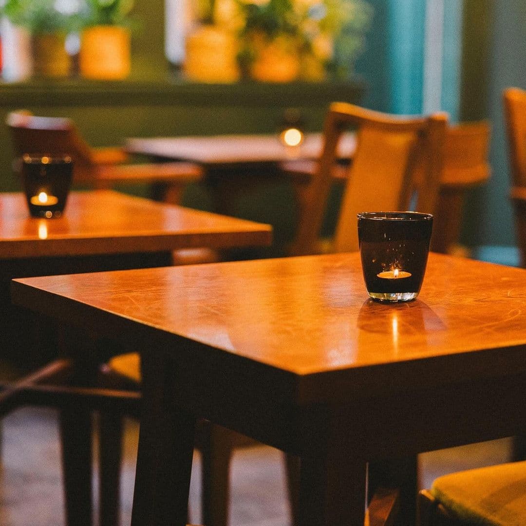 Holding Patterns cozy cafe interiors warm lighting tables A cozy interior scene at Holding Patterns, featuring wooden tables with a candle in a glass on one table. Soft lighting creates a warm atmosphere, with plants in the background adding a touch of greenery.