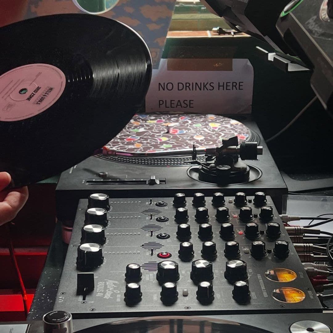Holding Patterns dj setup vinyl turntables music nightlife A close-up of a DJ's turntable setup at Holding Patterns. A hand holds a vinyl record above the turntable, while a colorful patterned platter is visible. The DJ mixer features various knobs and buttons, with a sign in the background that reads "NO DRINKS HERE PLEASE."