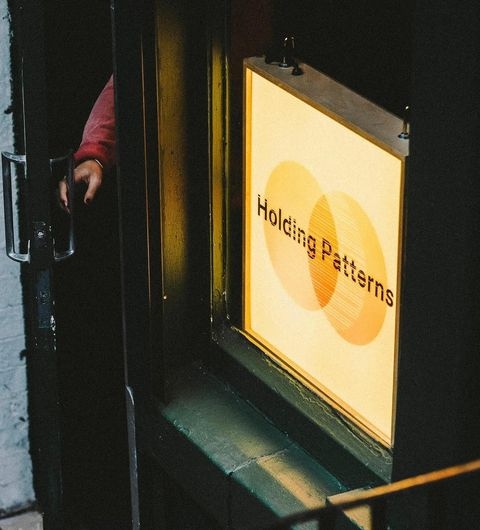 Holding Patterns sign entrance artistic light welcome A close-up view of a doorway with a sign that reads "Holding Patterns." The sign is illuminated, showcasing a modern design with overlapping shapes. A hand is seen reaching for the door, suggesting entry into the location.