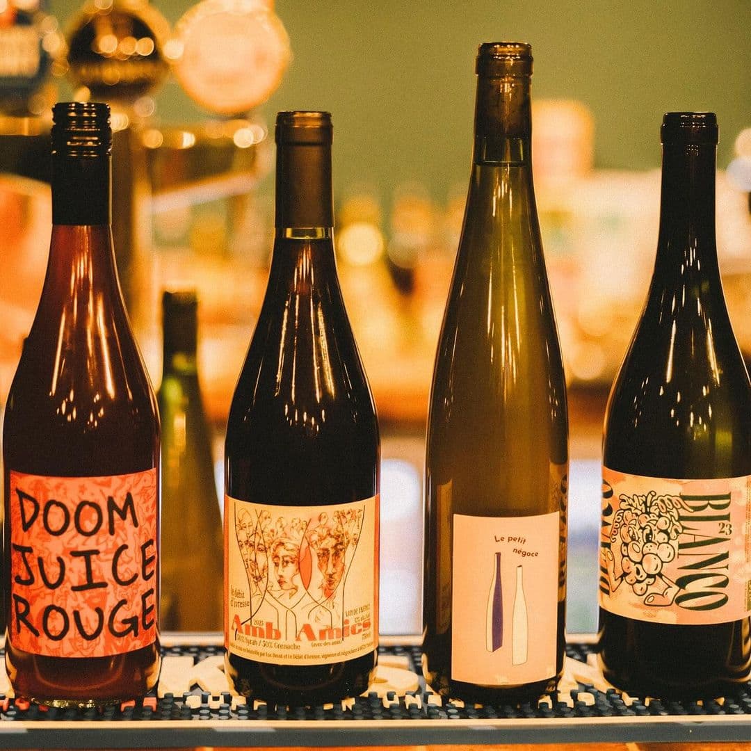 Holding Patterns wine bottles tasting tourism experience Four bottles of wine are displayed on a bar counter. The labels feature colorful designs, including "Doom Juice Rouge," a bottle with artistic illustrations, and "Le petit négociant." The background is softly blurred, highlighting the bottles in the foreground. This image represents the vibrant atmosphere at Holding Patterns, a destination for wine enthusiasts.