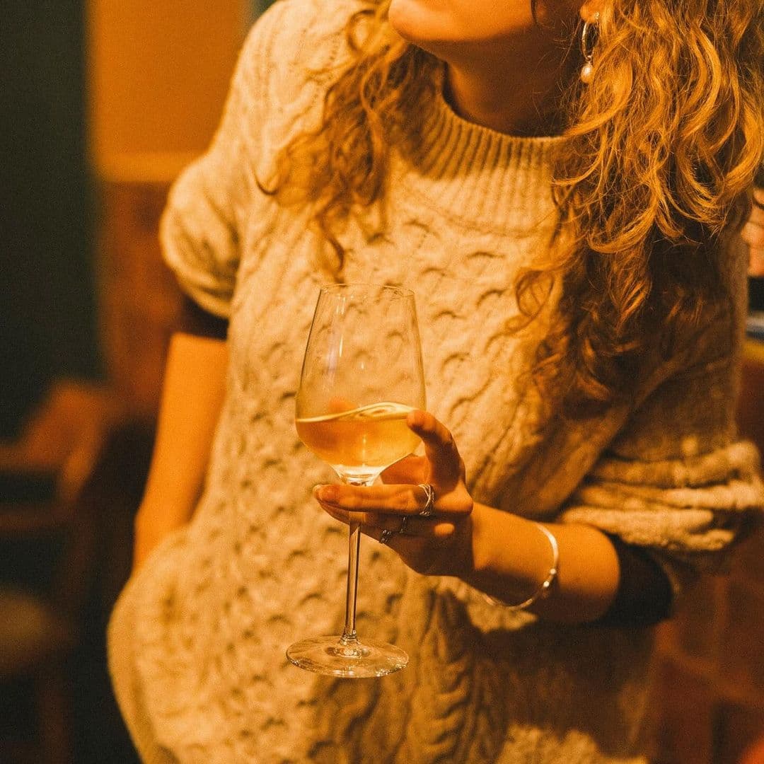 Holding Patterns wine glass relaxation evening vibes A person wearing a cozy, knitted sweater holds a glass of white wine, smiling softly in a warm, inviting atmosphere. The background features dim lighting, creating a relaxed ambiance. This image captures a moment of enjoyment at Holding Patterns.