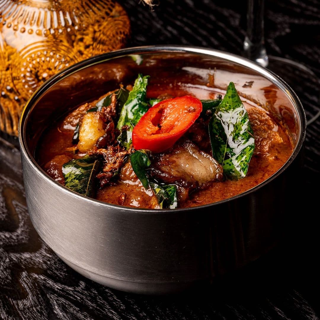 Kerala canteen authentic dish spicy flavors local cuisine A bowl of flavorful curry featuring tender meat, garnished with fresh green leaves and a slice of red chili. The dish is served in a stainless steel bowl, with a decorative traditional pot in the background. This is a delicious offering from Kerala canteen.