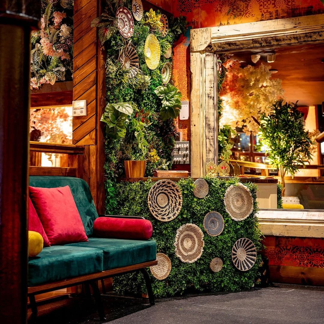 Kerala canteen cozy decor greenery artistic interior A cozy corner in Kerala Canteen featuring a green wall adorned with decorative plates and plants. A plush teal sofa with colorful cushions invites relaxation, while warm lighting creates a welcoming atmosphere.