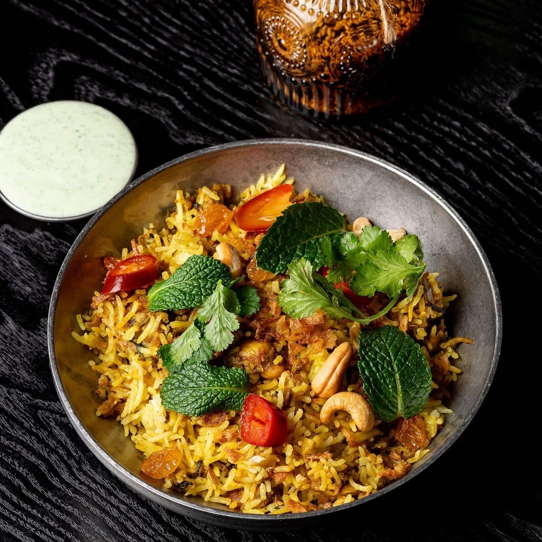 Kerala canteen delicious biryani authentic cuisine food A bowl of flavorful biryani topped with fresh mint leaves, coriander, and cherry tomatoes. The dish is accompanied by a small container of green sauce, set against a dark wooden background. This image represents the culinary delights at Kerala canteen.