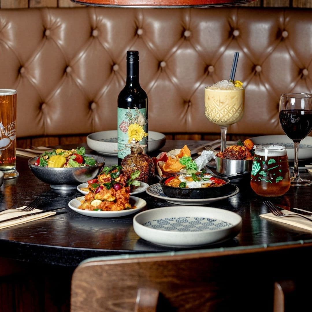 Kerala canteen delicious food dining experience beverages A beautifully arranged table at Kerala canteen featuring a variety of dishes, including colorful salads, spicy appetizers, and a selection of drinks. The setting includes a bottle of wine, a cocktail garnished with fruit, and a glass of beer. The backdrop is a stylish leather booth, creating a warm and inviting atmosphere for dining.