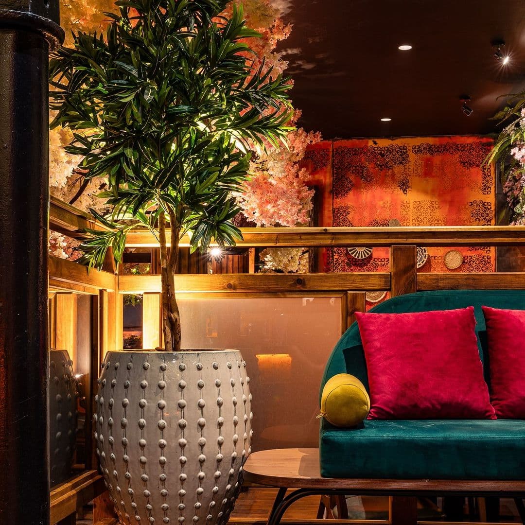 Kerala canteen interior decor cozy atmosphere modern design Cozy interior of Kerala Canteen featuring a green sofa with pink cushions, a decorative pot with a leafy plant, and warm ambient lighting. The background showcases colorful wall art and wooden accents, creating a welcoming atmosphere.