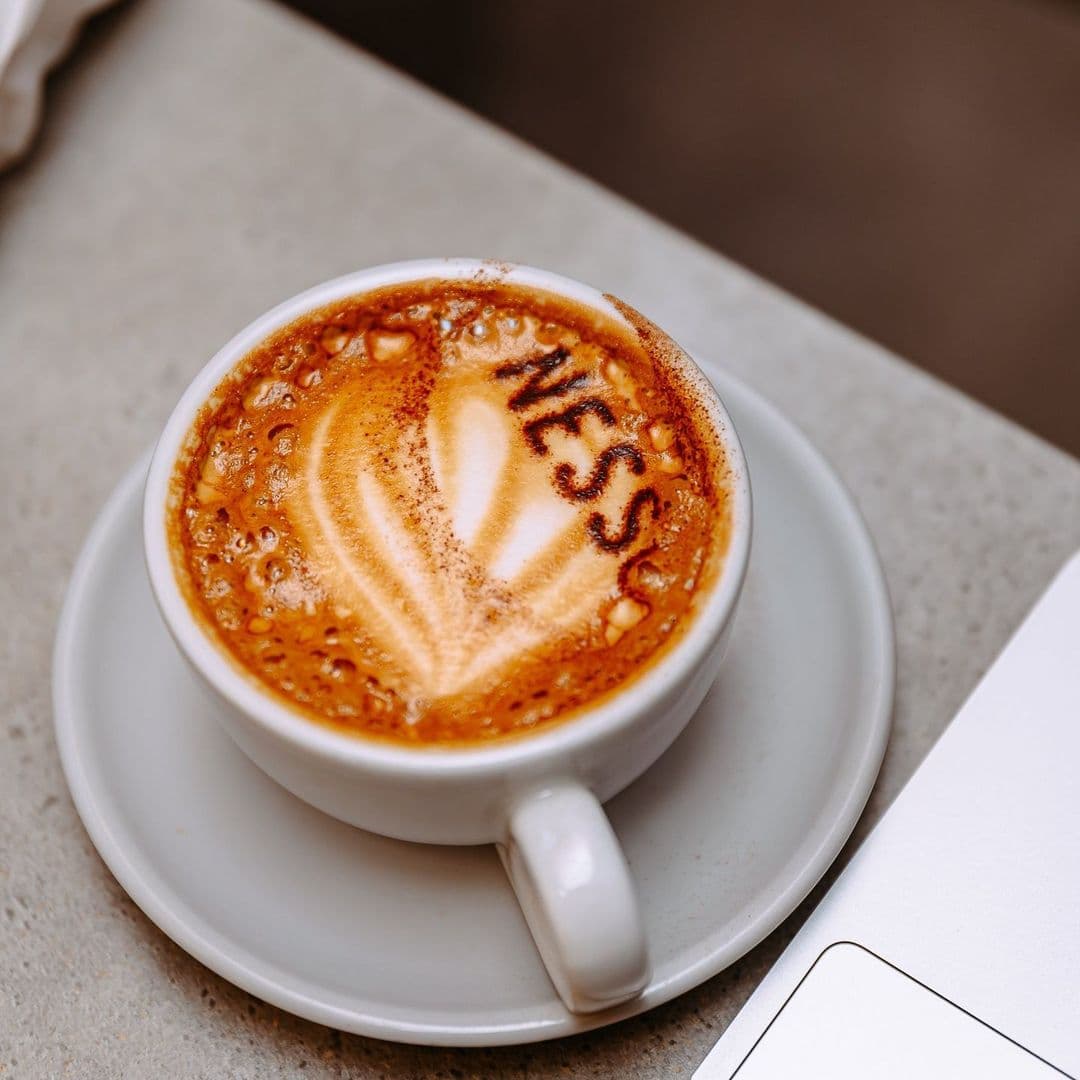 Nesso Coffee artisan coffee cup latte art cafe A close-up of a latte in a white cup with the word "NESS" artfully crafted in the foam. The cup sits on a small saucer on a light-colored table, with a glimpse of a laptop nearby. This image captures the cozy atmosphere of Nesso Coffee.