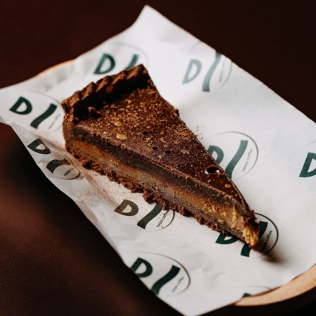 Nesso Coffee chocolate tart dessert gourmet bakery A slice of rich chocolate tart on branded paper, served at Nesso Coffee. The tart features a dark chocolate layer atop a golden filling, with a crumbly crust.