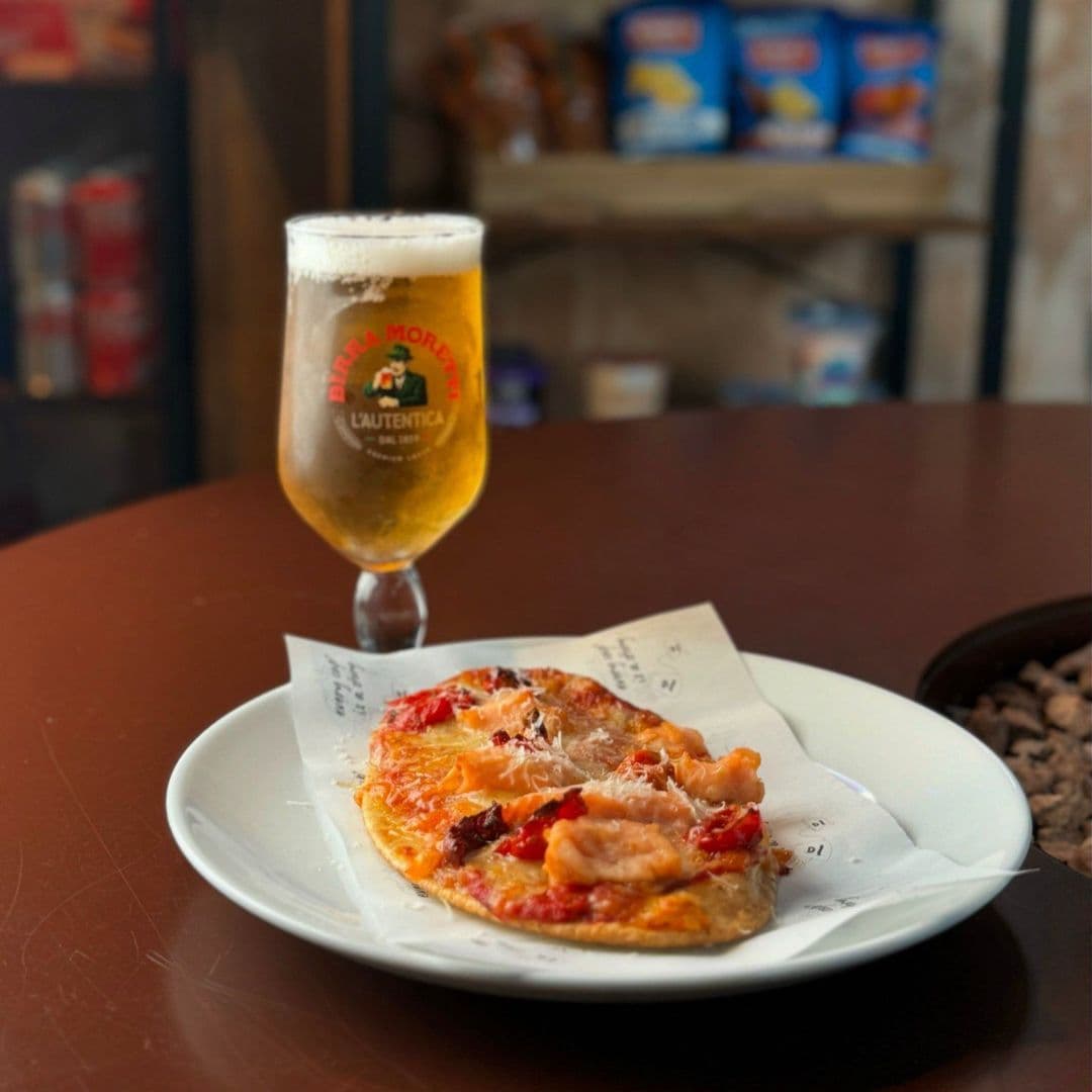 Nesso Coffee delicious food beer local cuisine A plate of delicious pizza topped with tomatoes and cheese sits on a wooden table, accompanied by a glass of beer. The setting is warm and inviting, featuring a rustic background with shelves of snacks. This image represents the dining experience at Nesso Coffee.