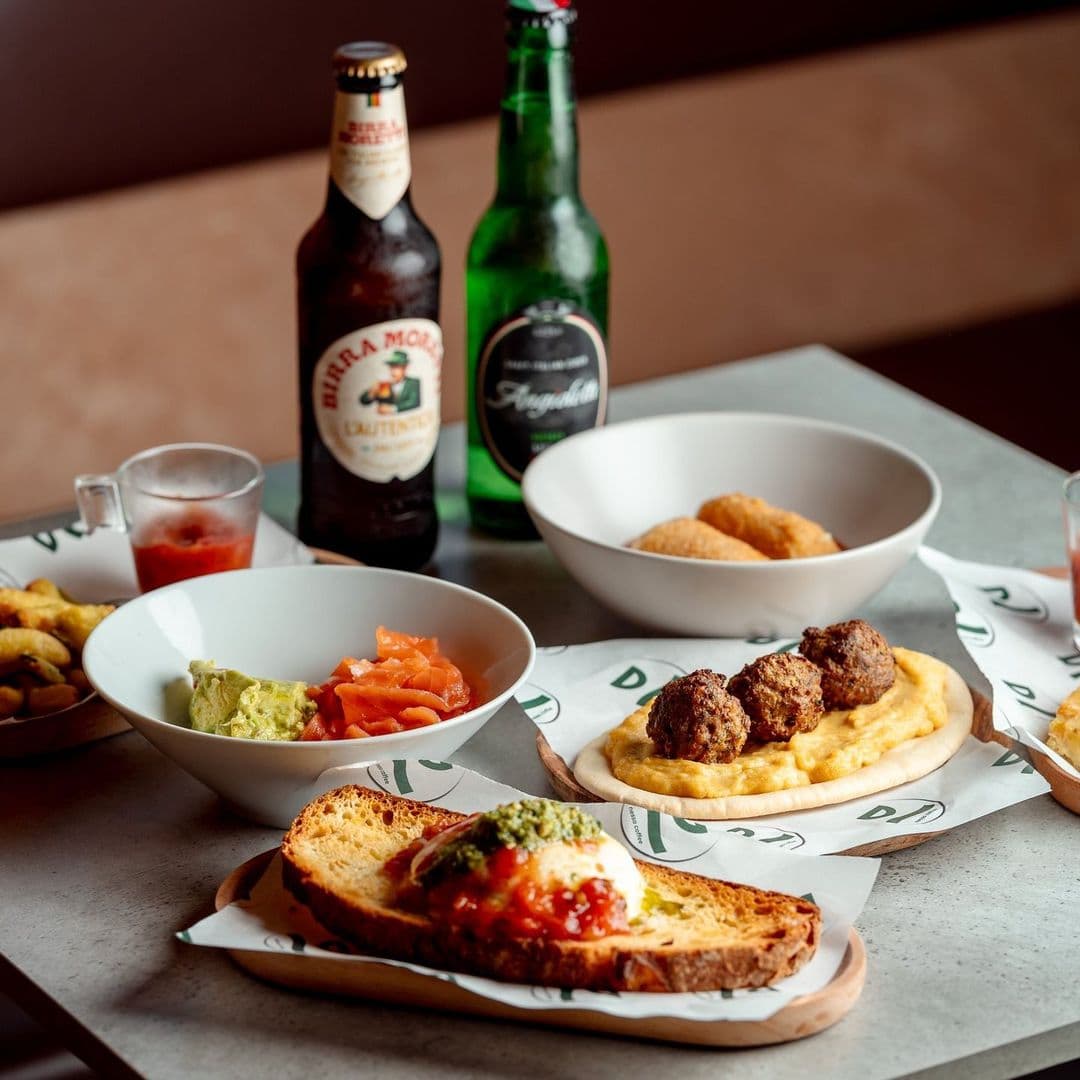 Nesso Coffee delicious food beverages gourmet experience A delicious spread of food and drinks at Nesso Coffee. The table features a variety of dishes, including a slice of bread topped with sauce and herbs, a bowl of guacamole, smoked salmon, and meatballs served on a creamy base. Two bottles of beer and a shot glass are also visible, along with a bowl of fried snacks. The setting is warm and inviting, perfect for enjoying a meal.