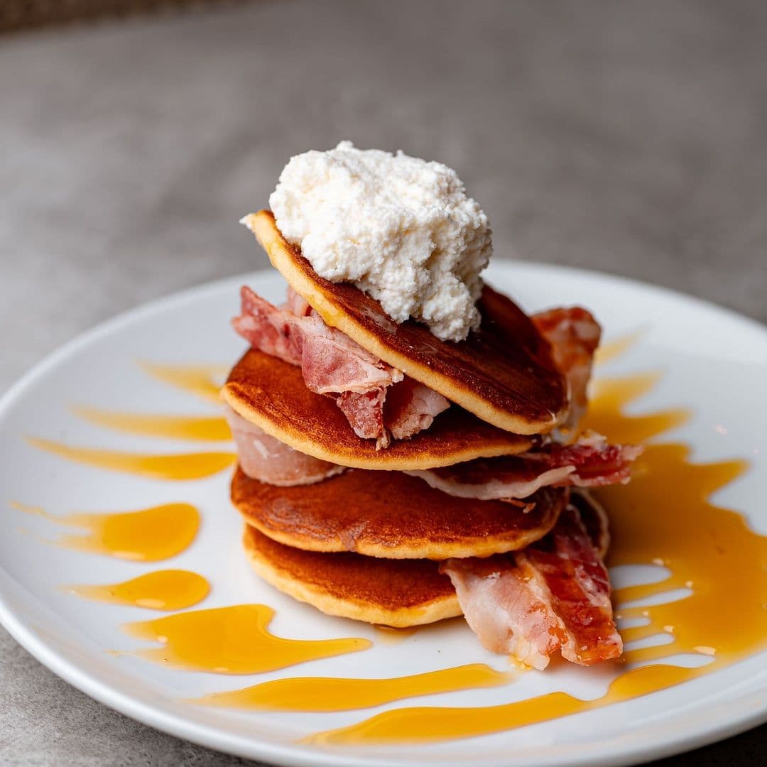 Nesso Coffee pancakes bacon maple syrup breakfast Delicious stack of pancakes topped with whipped cream and crispy bacon, drizzled with syrup, served on a white plate at Nesso Coffee.