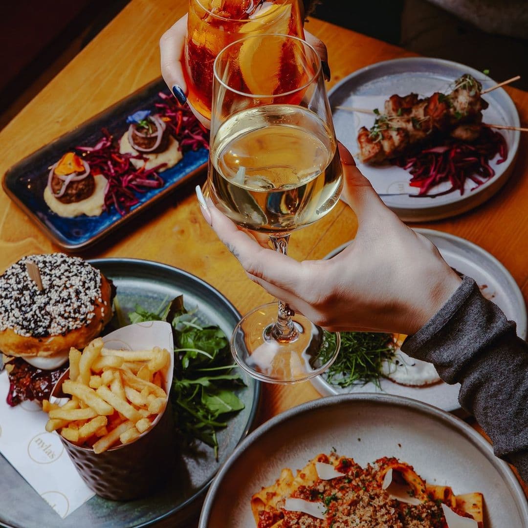 Nyla s dining experience gourmet food drinks socializing A beautifully arranged table at Nyla’s featuring a variety of dishes. In the foreground, a hand holds a glass of white wine, while another hand raises a colorful cocktail. Plates include a burger topped with sesame seeds, crispy French fries, and a dish of pasta with garnishes. The setting is warm and inviting, perfect for enjoying a meal with friends.