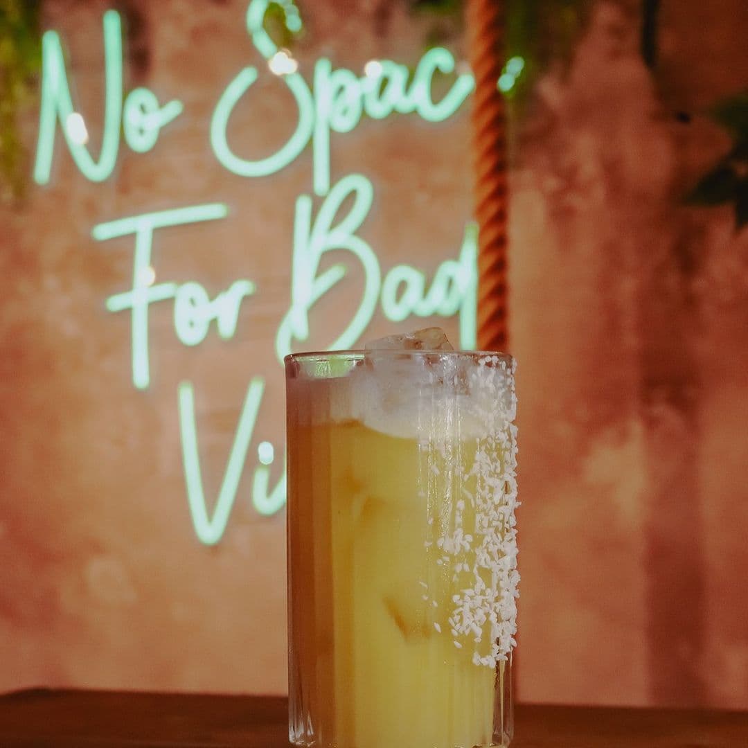 Nyla s tropical cocktail refreshing drink vibrant background A refreshing cocktail with a frothy top and a salted rim sits on a wooden table. In the background, a neon sign reads "No Space For Bad Vibes." This vibrant scene captures the lively atmosphere at Nyla’s.