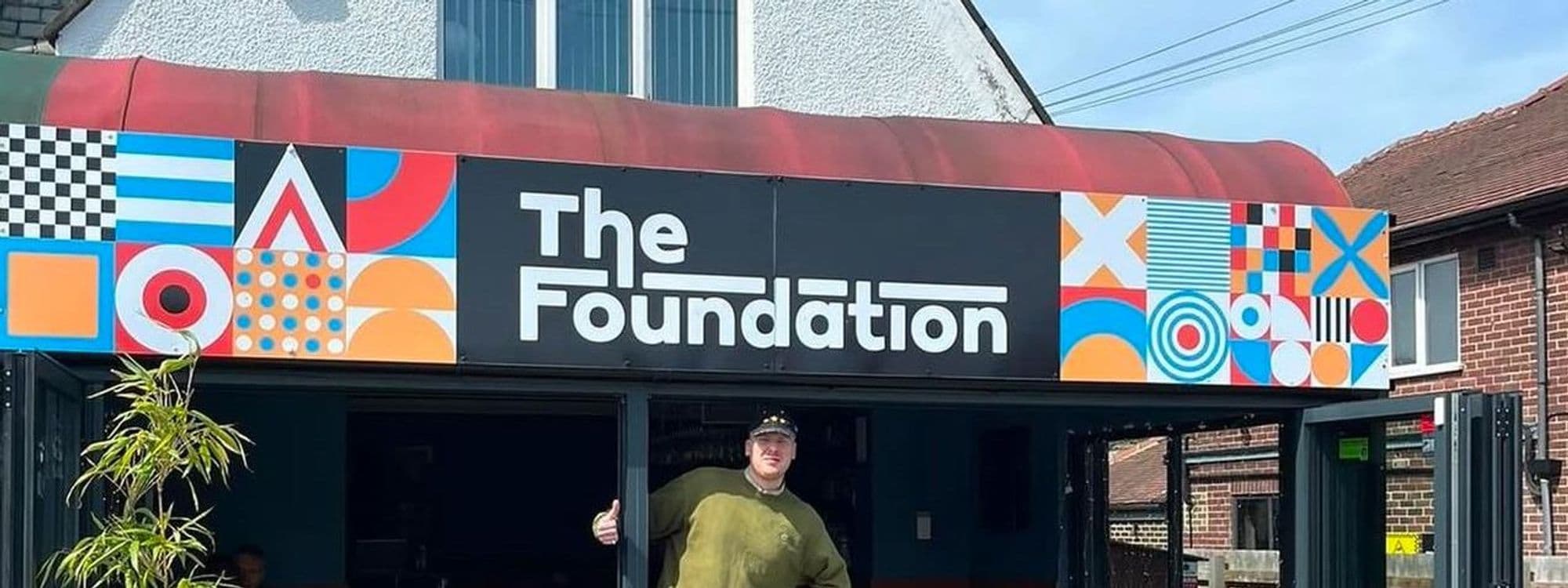 A person stands in front of a colorful entrance to The Foundation, featuring a vibrant mural with geometric patterns and bold colors. The sign prominently displays the name "The Foundation." The setting is bright and inviting, with greenery visible nearby.