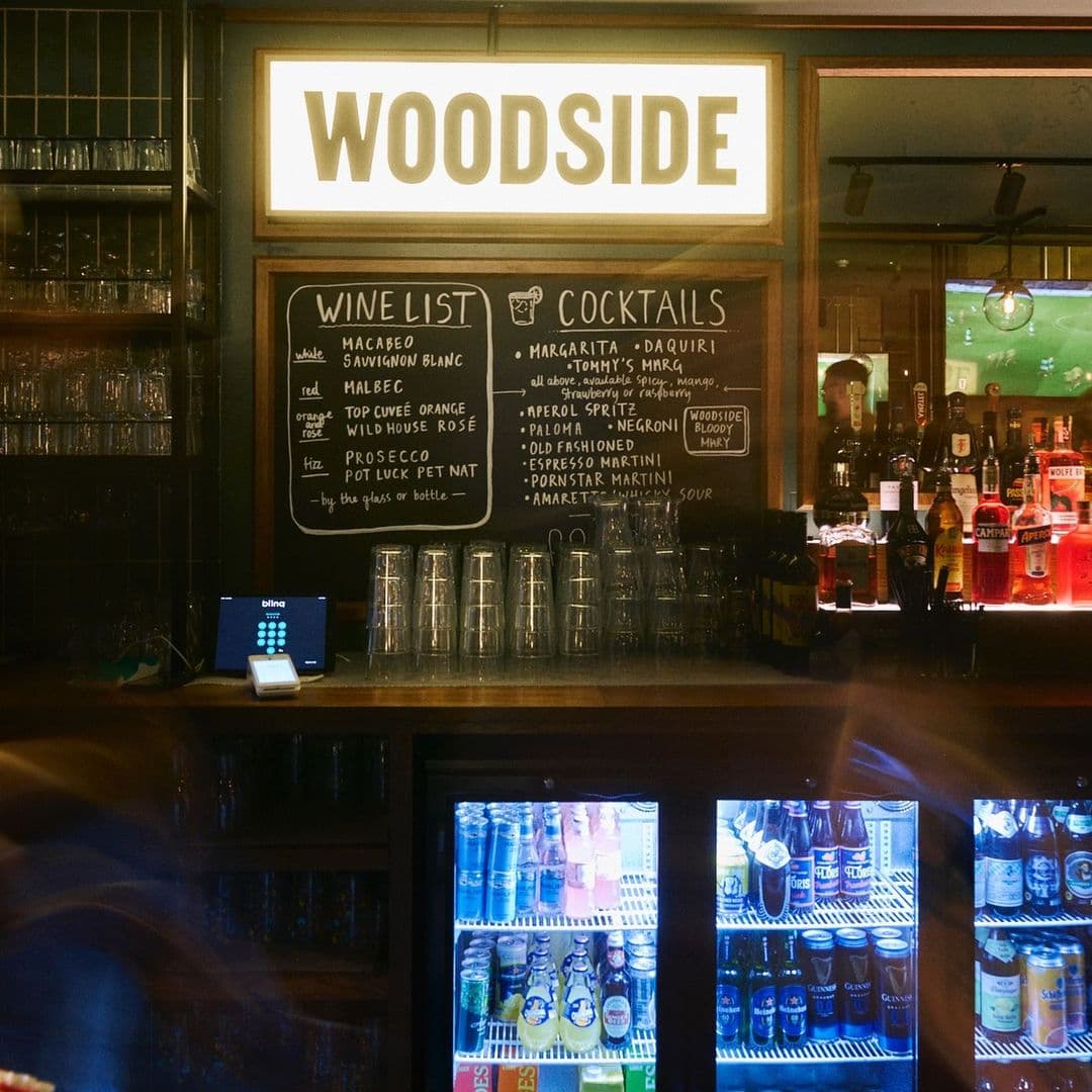 Woodside Neighbourhood Bar Eatery bar drinks menu cocktails relaxation nightlife Alt text: Interior view of Woodside Neighbourhood Bar & Eatery, featuring a wooden bar with a wine and cocktail menu displayed on a chalkboard. Glasses are stacked on the bar, and a refrigerator showcases various drinks. The bar has a warm, inviting atmosphere.