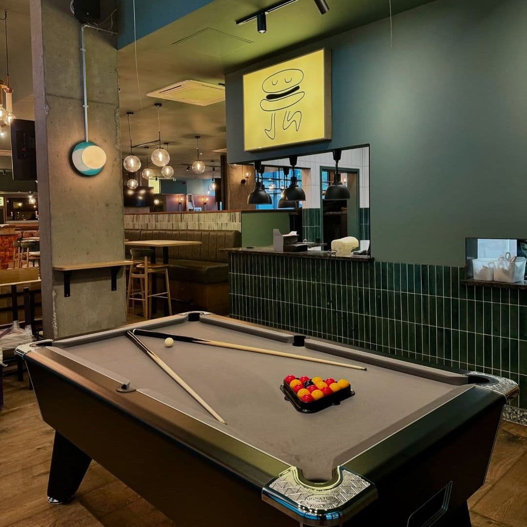Woodside Neighbourhood Bar Eatery billiards game interior casual dining ambiance A stylish interior of Woodside Neighbourhood Bar & Eatery featuring a pool table with colorful balls arranged for play. The space is illuminated by hanging lights and has a modern design with green walls and wooden accents. A playful burger-themed artwork is displayed on the wall, adding a fun touch to the atmosphere.
