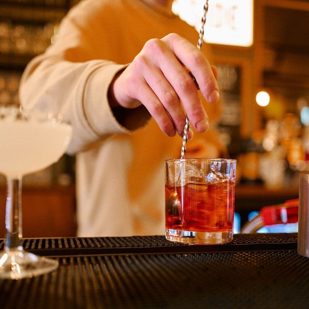 Woodside Neighbourhood Bar Eatery cocktail preparation bartender drink mixology A bartender stirs a cocktail in a glass at Woodside Neighbourhood Bar & Eatery. In the foreground, a second glass with a clear drink is visible. The background shows a cozy bar setting with warm lighting.