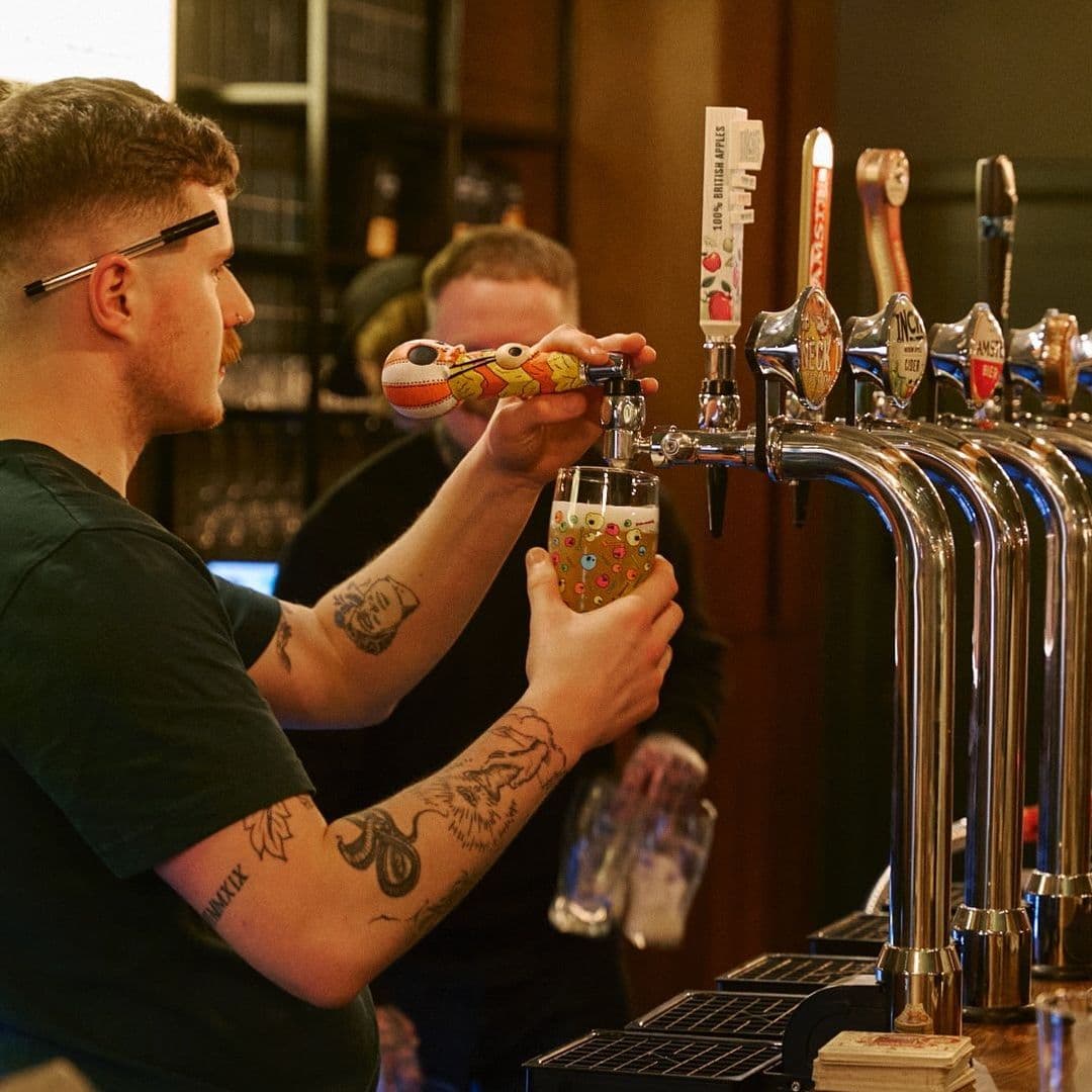Woodside Neighbourhood Bar Eatery craft beer tap bartender drinks social A bartender at Woodside Neighbourhood Bar & Eatery pours a drink from a colorful tap while holding a glass decorated with playful designs. In the background, another person is visible, adding to the lively atmosphere of the bar.