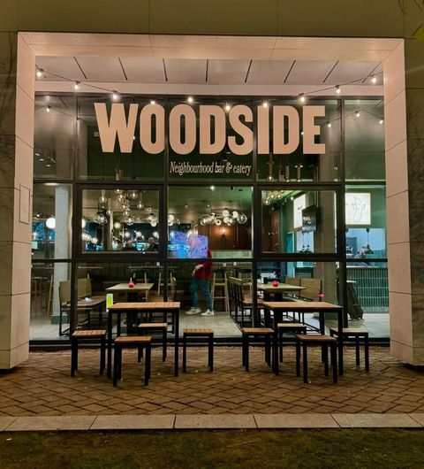 Woodside Neighbourhood Bar Eatery exterior night lights dining Exterior view of Woodside Neighbourhood Bar & Eatery, featuring large windows with the name "WOODSIDE" prominently displayed. The entrance has outdoor seating with tables and chairs, and warm lighting creates an inviting atmosphere. Inside, the bar is visible with stylish decor and a cozy ambiance.