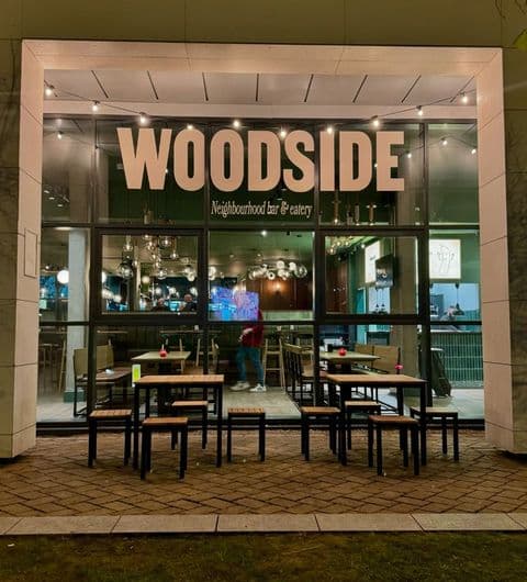 Woodside Neighbourhood Bar Eatery exterior night lights dining Exterior view of Woodside Neighbourhood Bar & Eatery, featuring large windows with the name "WOODSIDE" prominently displayed. The entrance has outdoor seating with tables and chairs, and warm lighting creates an inviting atmosphere. Inside, the bar is visible with stylish decor and a cozy ambiance.