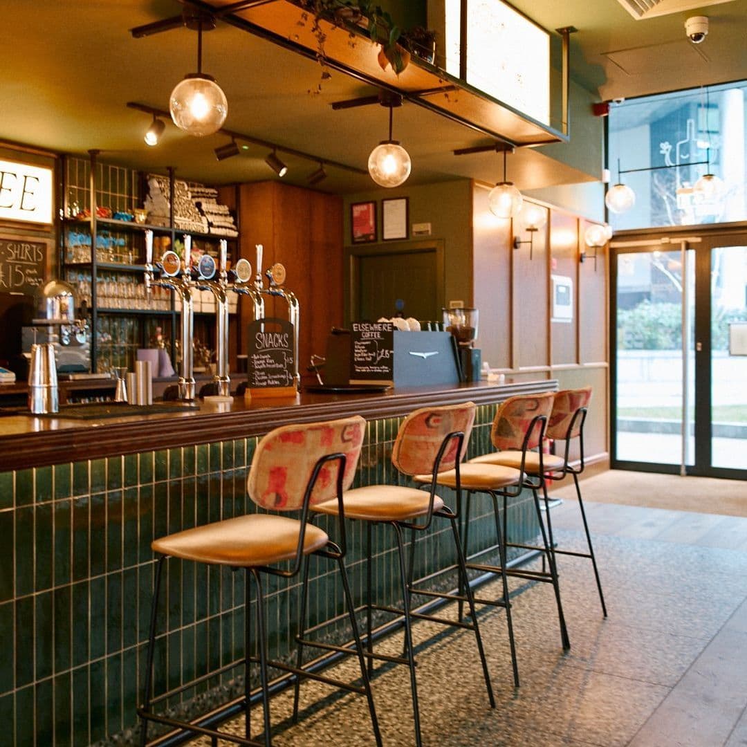 Woodside Neighbourhood Bar Eatery modern bar interior cozy seating lighting Alt text: Interior of Woodside Neighbourhood Bar & Eatery featuring a wooden bar with several beer taps, stylish bar stools, and soft lighting. The space has a warm, inviting atmosphere with green tiles and a large window letting in natural light.