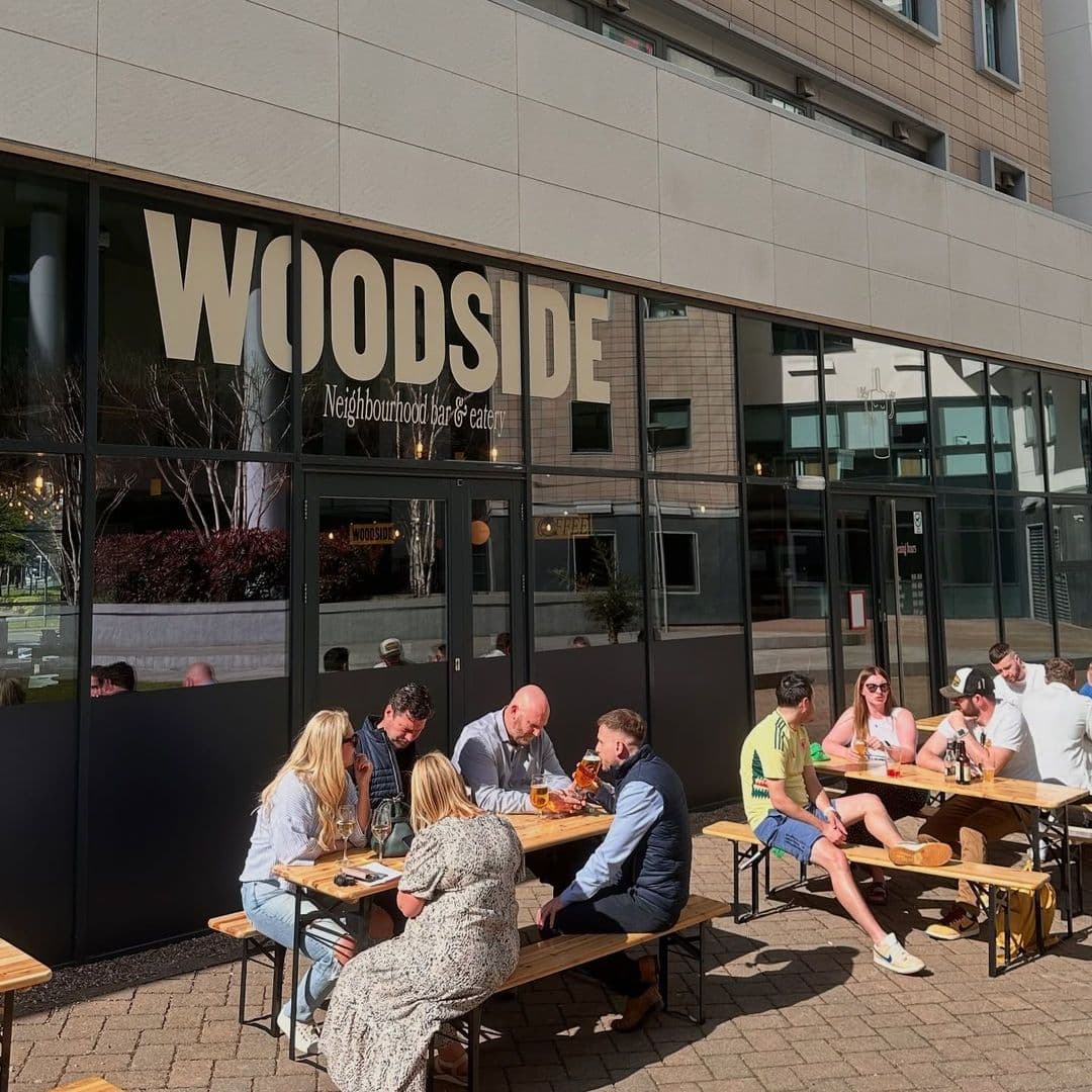 Woodside Neighbourhood Bar Eatery outdoor dining happy customers sunny day relaxation People enjoying drinks and meals at outdoor tables in front of Woodside Neighbourhood Bar & Eatery. The setting features a modern building with large windows and a welcoming atmosphere.