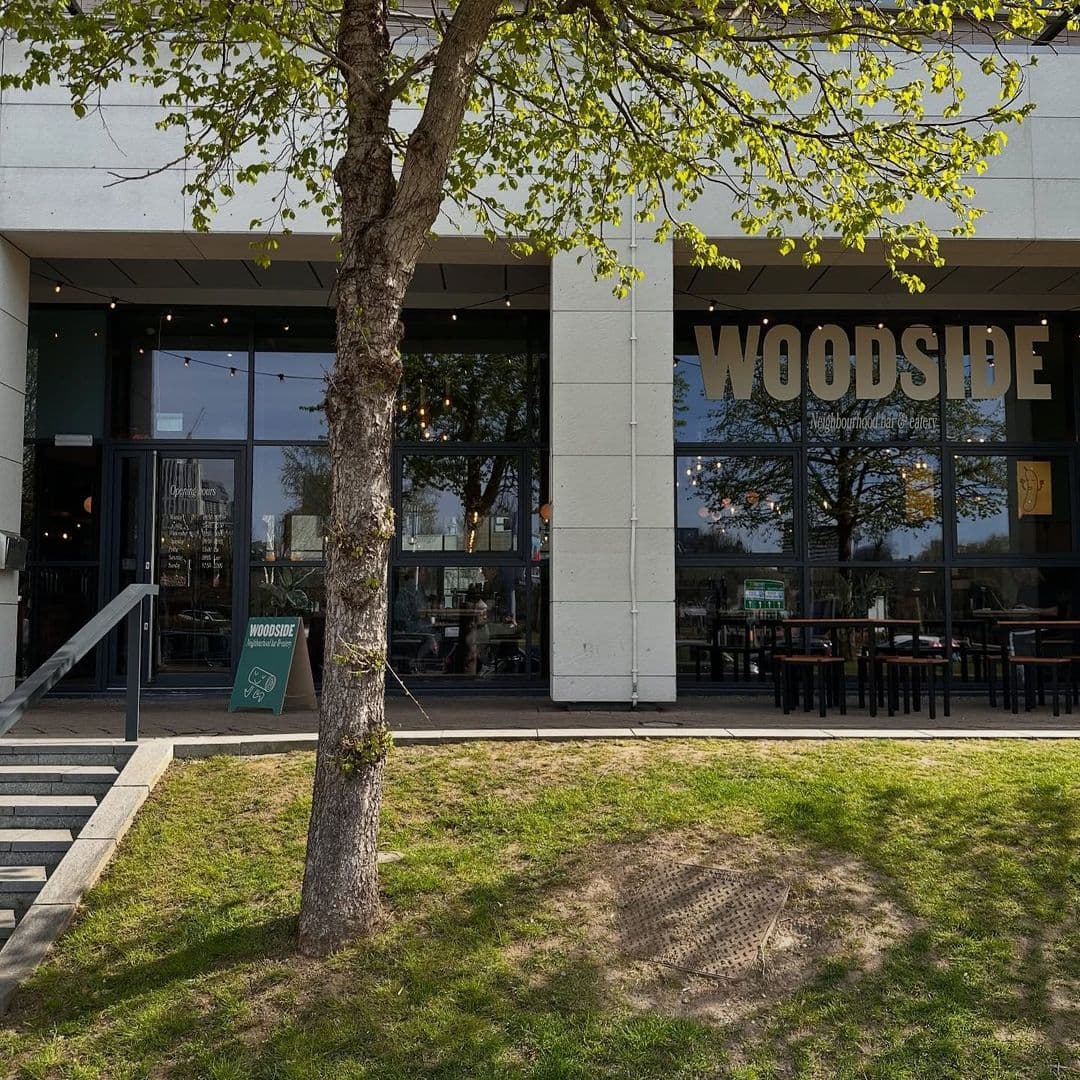 Woodside Neighbourhood Bar Eatery outdoor dining modern architecture relaxing atmosphere Exterior view of Woodside Neighbourhood Bar & Eatery, featuring a modern building with large windows, a welcoming entrance, and outdoor seating. A small tree and green grass are visible in front, creating a cozy atmosphere.