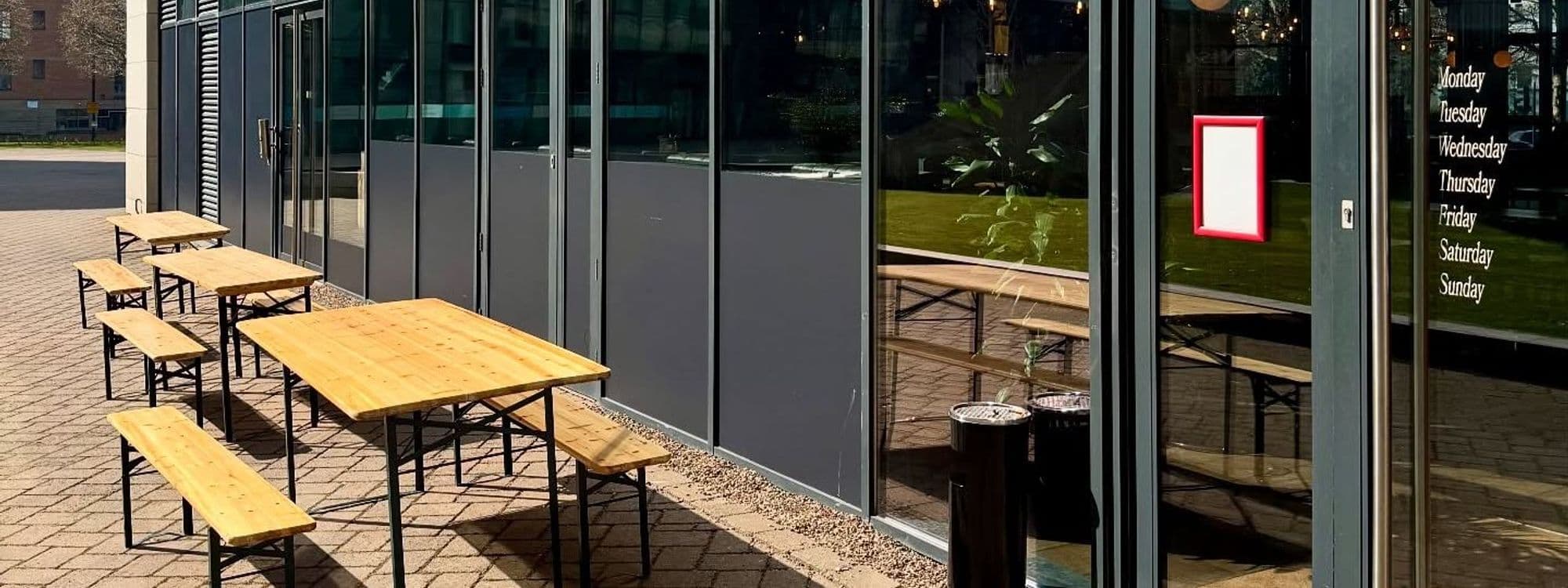 Outdoor seating area at Woodside Neighbourhood Bar & Eatery, featuring wooden tables and benches on a paved patio. Large glass windows provide a view of the interior.