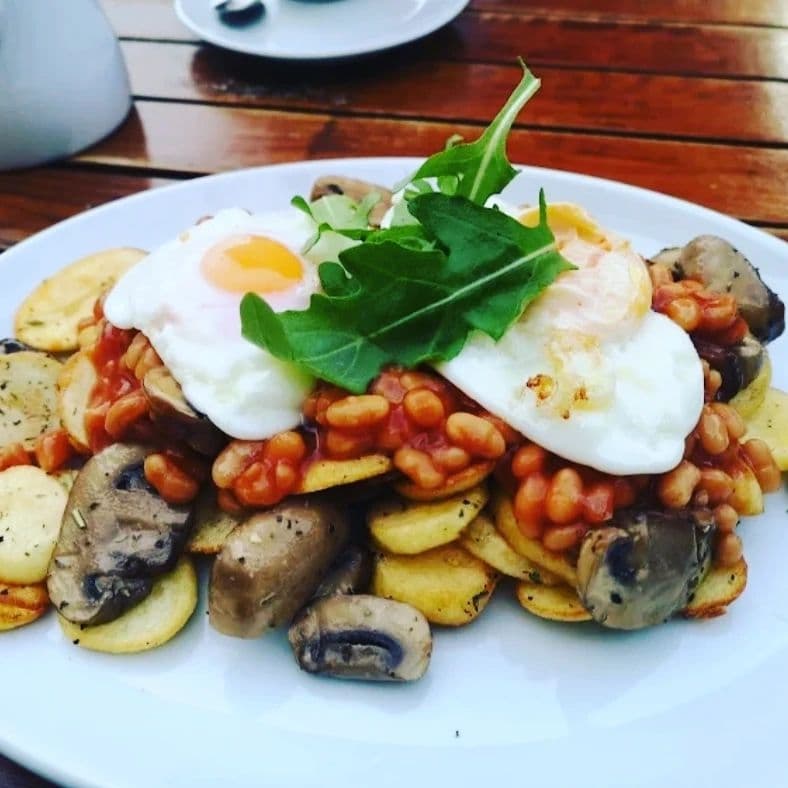 Delicious Breakfast Stack with Poached Eggs A hearty plate of sautéed potatoes topped with baked beans, mushrooms, poached eggs, and fresh arugula, set on a wooden table.