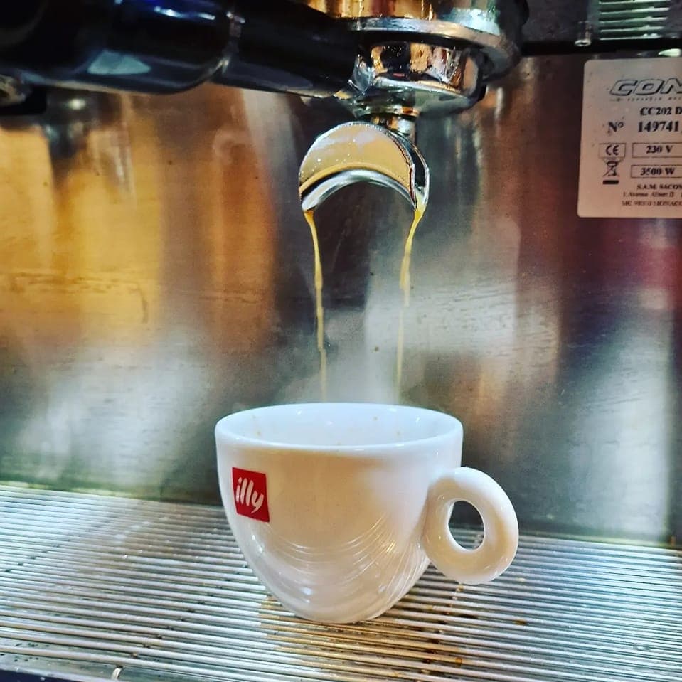Freshly Brewed Espresso Pouring Rich espresso drips from a shiny machine into a white cup with a red logo, set against a metallic backdrop.