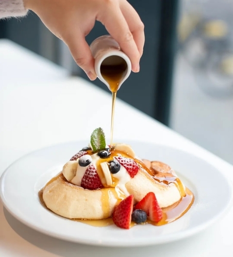 Decadent Pancake Stack with Fresh Berries A hand drizzles syrup over a fluffy pancake topped with strawberries, blueberries, and a mint leaf on a white plate.