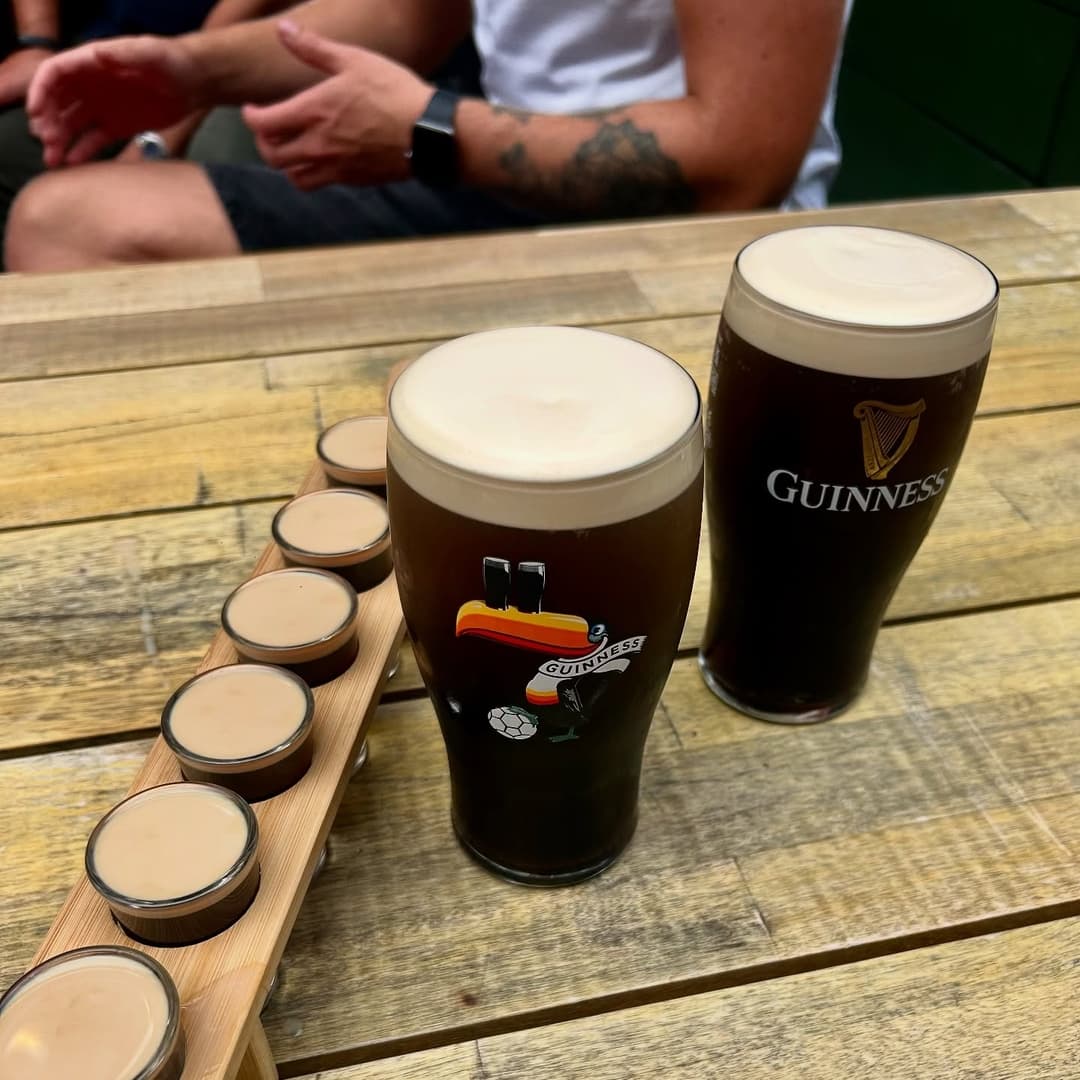 Guinness Pints with Tasting Flight Two pints of Guinness sit on a wooden table alongside a flight of creamy shot glasses, creating a lively pub atmosphere.