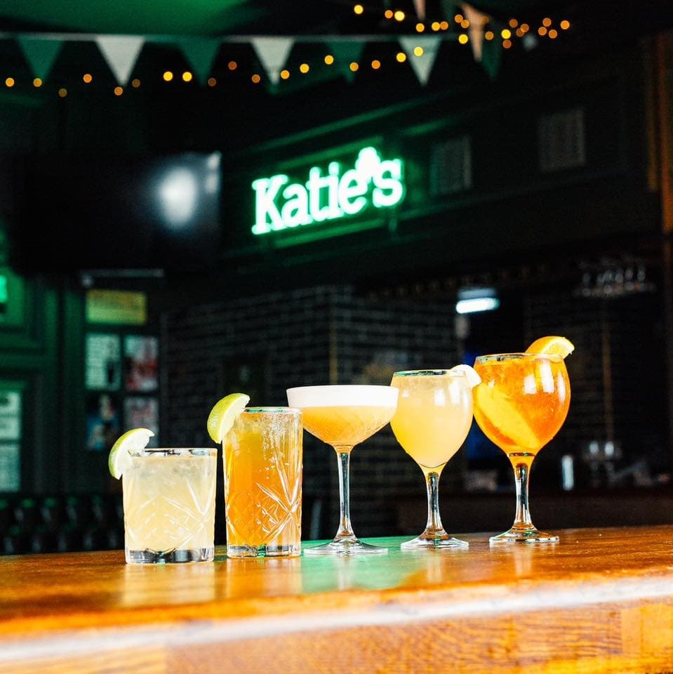 Vibrant Selection of Signature Cocktails Five colorful cocktails with garnishes line a wooden bar, illuminated by soft lights and a glowing "Katie's" sign in the background.
