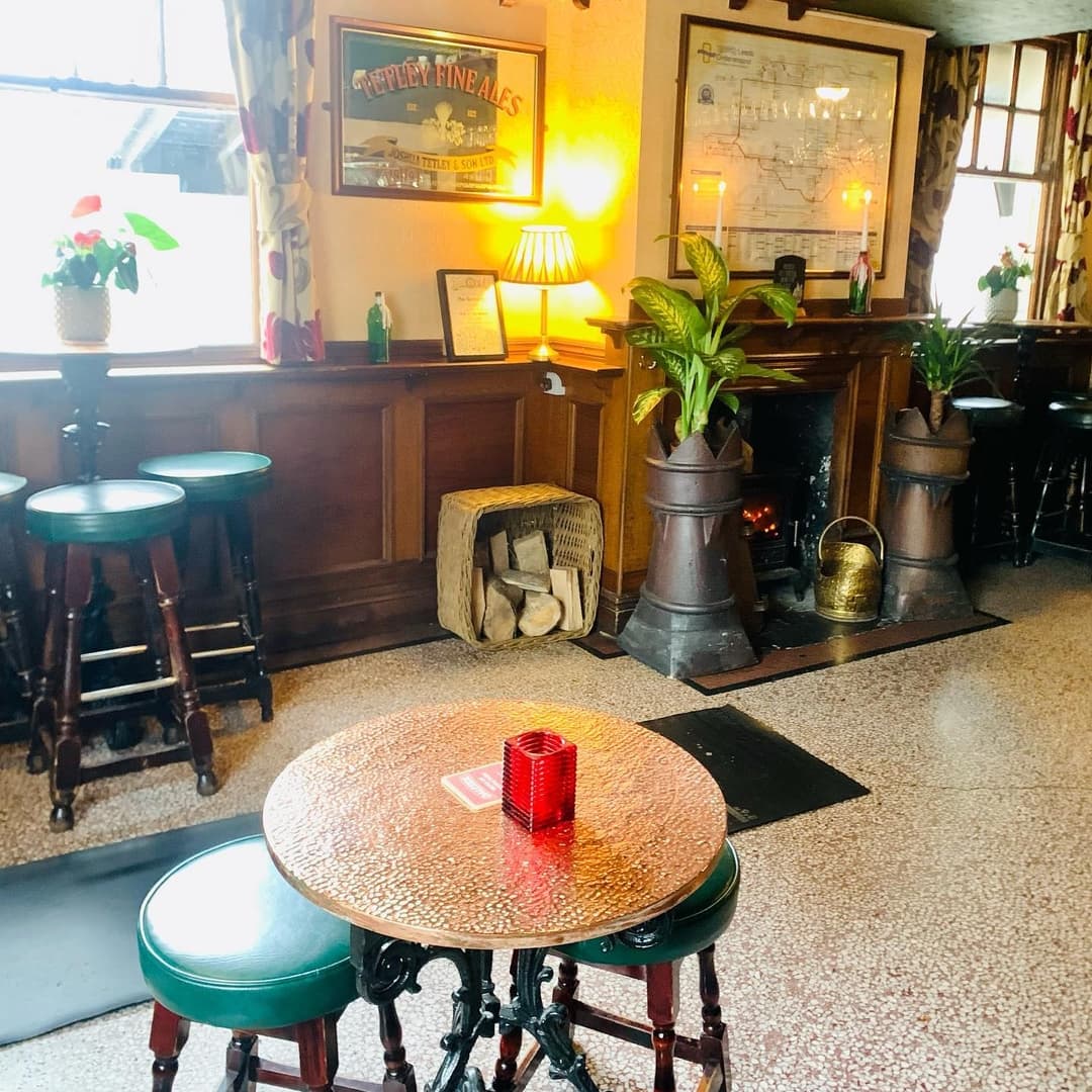 Cozy Pub Corner with Rustic Charm Cozy pub interior with wooden bar, green stools, a glowing fireplace, and potted plants, creating a warm atmosphere.