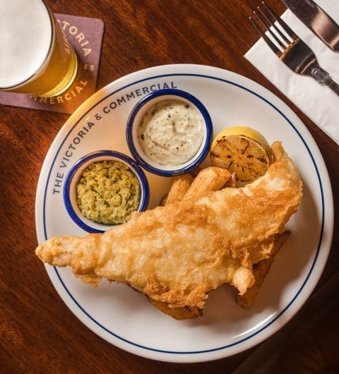 the-victoria-and-commercial-beer-battered-fish-chips Beer-battered fish with tartare sauce, mushy peas and lemon on a branded Victoria and Commercial plate with a pint of lager in the background