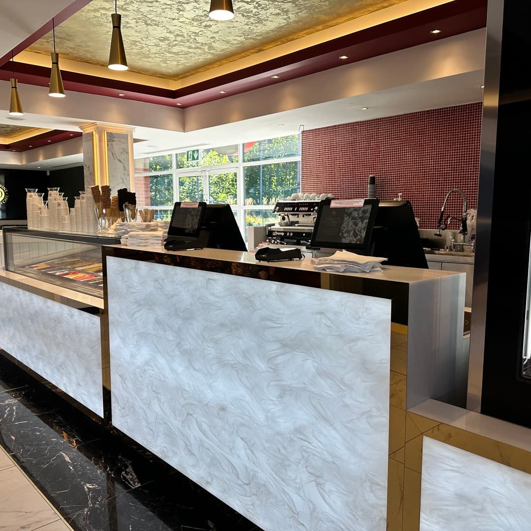 39 Desserts — counter with red mosaic feature wall The interior counter at 39 Desserts, Leeds, with marbled white panels, gold trim, hanging gold pendant lights, gold metallic ceiling and a red glass-mosaic feature wall behind.