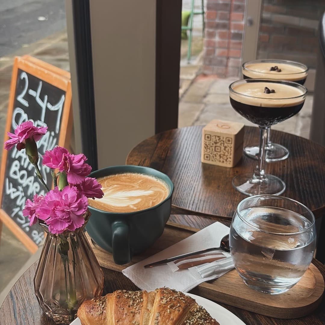 Ankave — coffee and cocktails table A flat white with latte art, a sesame croissant, two espresso martinis, and a vase of pink carnations on a wooden table at Ankave Coffee and Cocktails, Leeds.