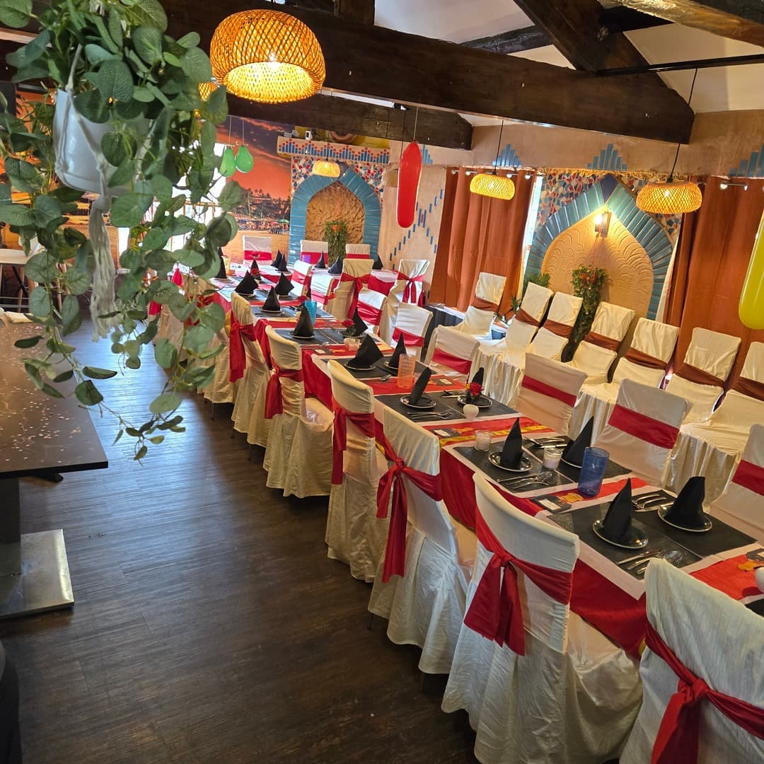 Bombay Junction — dining room with archway murals The Bombay Junction dining room in Bramley, Leeds, set with red sashes on white chair covers, exposed timber beams, hanging eucalyptus, woven pendant lights and Mughal-style archway murals on the back wall.
