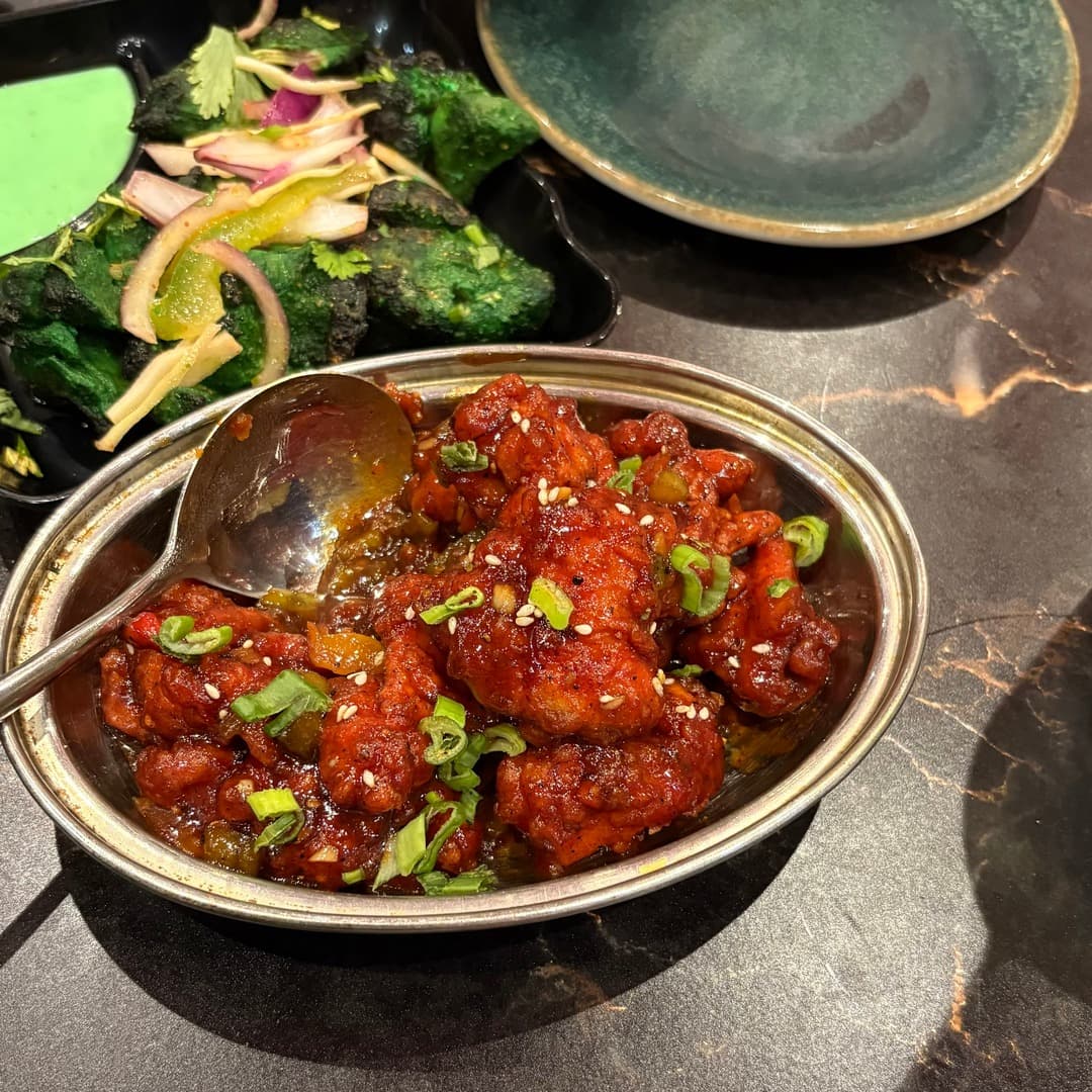 Bombay Junction — Indo-Chinese chicken Manchurian An Indo-Chinese chicken Manchurian at Bombay Junction, Leeds, in a metal serving dish with sliced spring onions and sesame seeds, alongside a side dish of green spinach pakora.