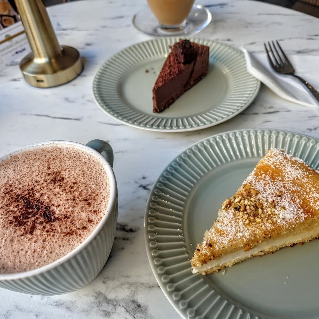 Jasmine Cafe — hot chocolate, cakes and karak tea A spiced hot chocolate, two slices of cake (chocolate and a coconut-pistachio sponge), and a karak tea on a marble table at Jasmine Cafe, Leeds.